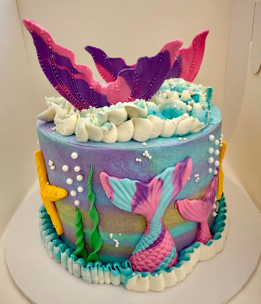 Colorful mermaid-themed cake with sea-themed decorations on a white surface.