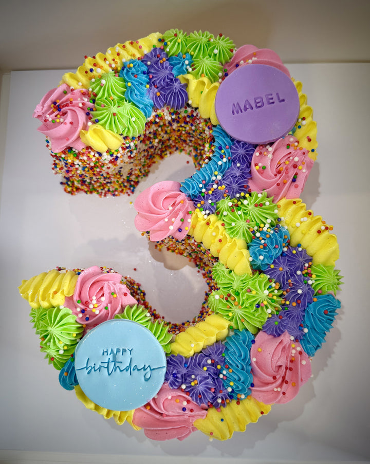 Colorful birthday cake shaped like the number '3' with frosting and a 'Mabel' button.