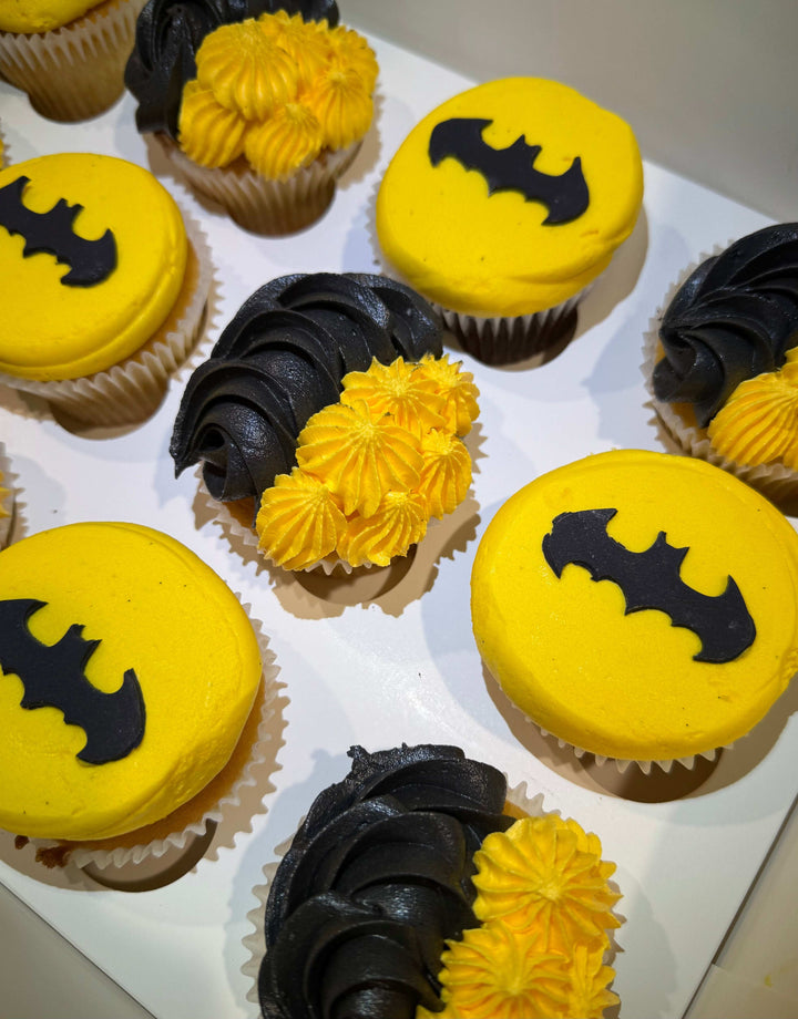 Cupcakes with yellow and black icing, featuring a bat symbol, on a white tray.