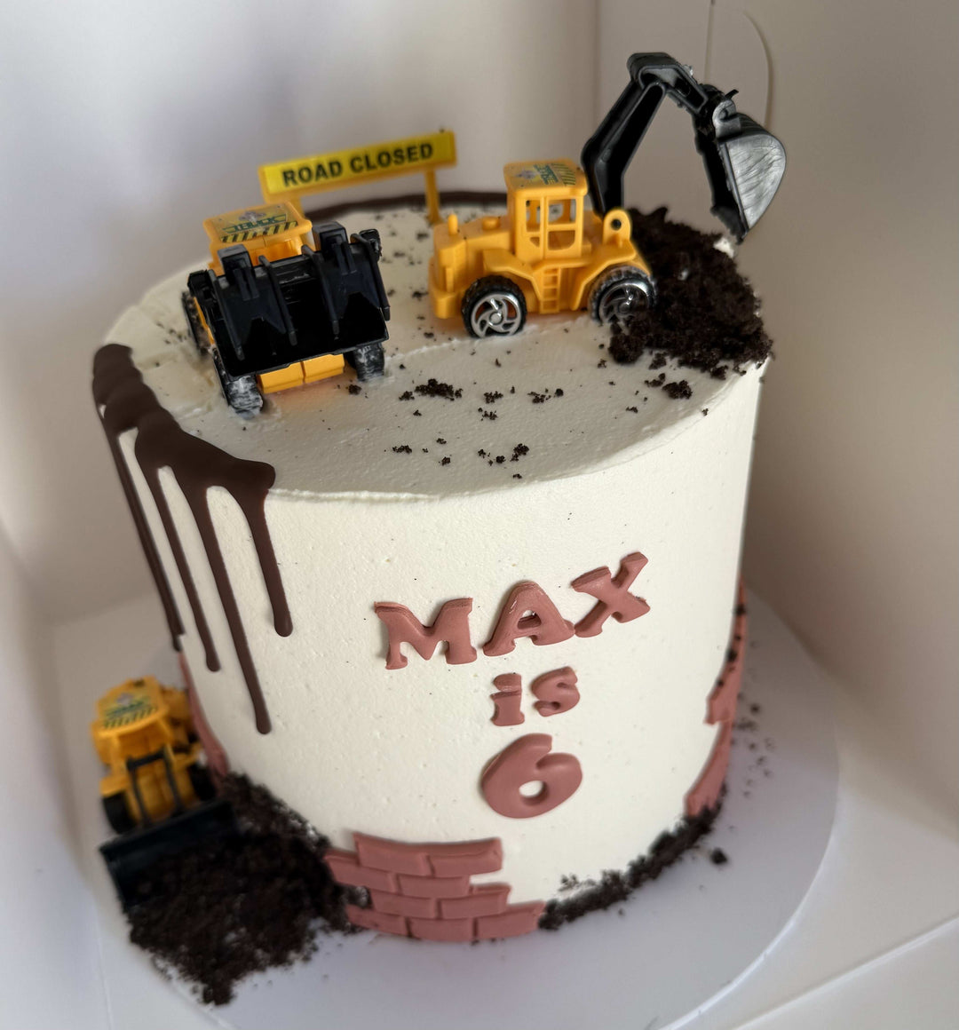 Construction-themed birthday cake with toy trucks and 'MAX is 6' text on a white background.