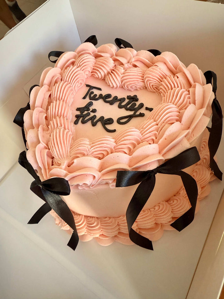 Pink cake with 'Twenty-fine' written on it, surrounded by black ribbons, on a white background.