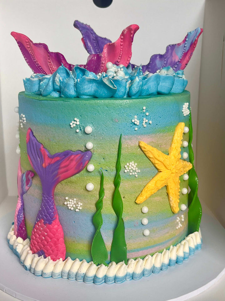 Colorful cake with mermaid tails, starfish, and seaweed decorations on a white background