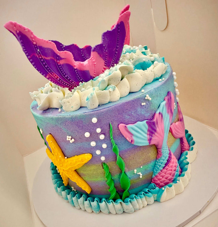 Colorful mermaid-themed cake with ocean-themed decorations on a white surface.